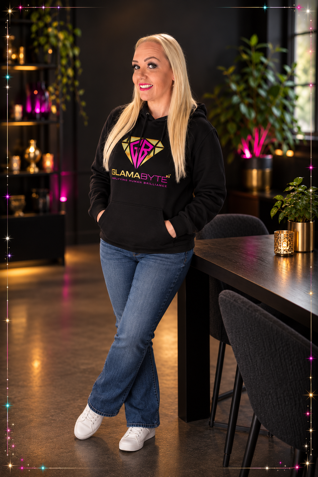 Smiling blonde woman in a black “GLAMA BYTE” hoodie stands beside a dining table in a warmly lit room.