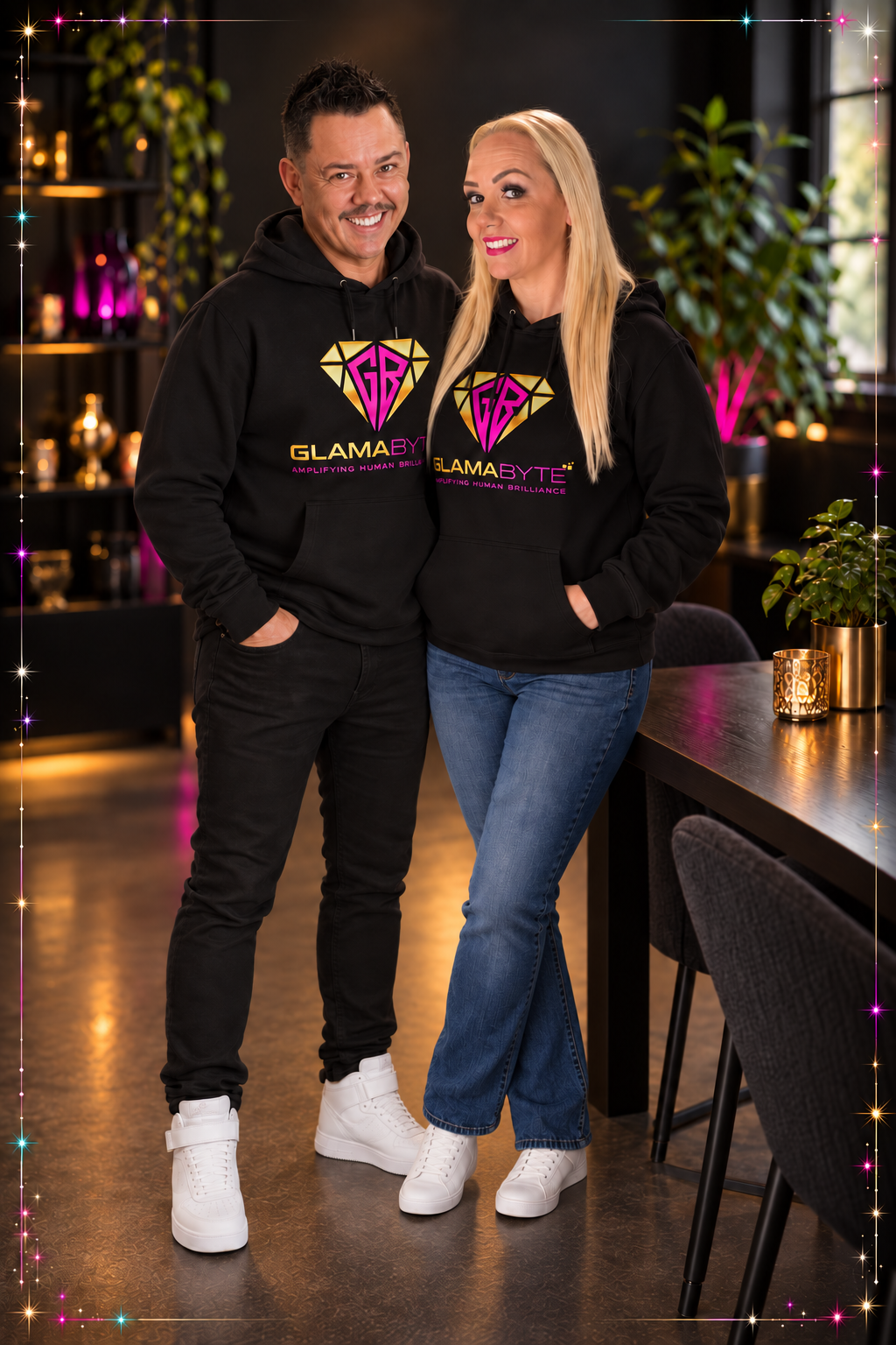 Smiling man and woman in matching black GLAMABYTE hoodies stand in a warmly lit lounge.